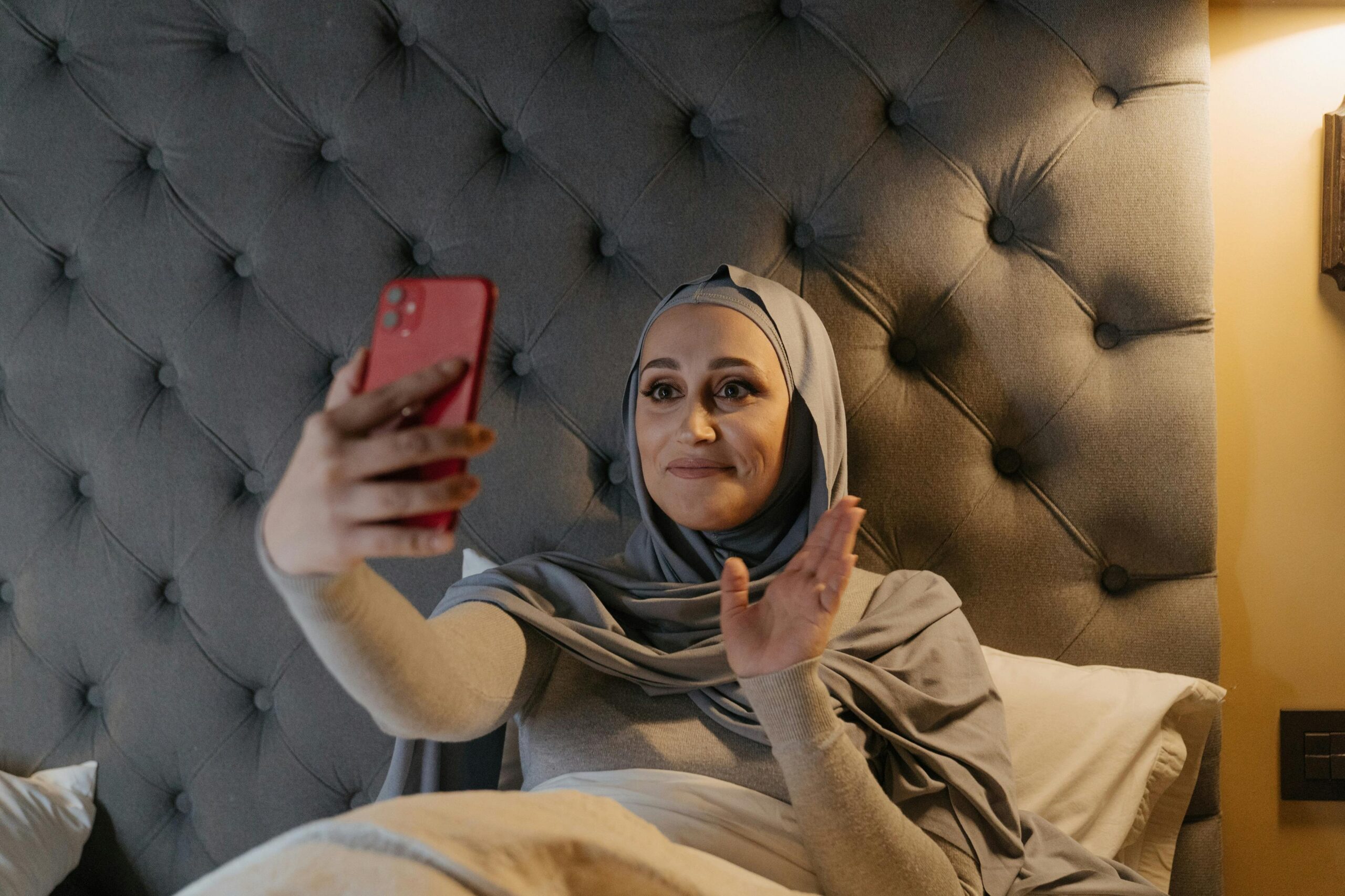 A woman in traditional hijab sits in bed, waving during a video call with a smartphone.